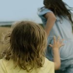 Two girls play chase in the grass, one younger wearing yellow, one older wearing blue, with a pale blue sky behind them.