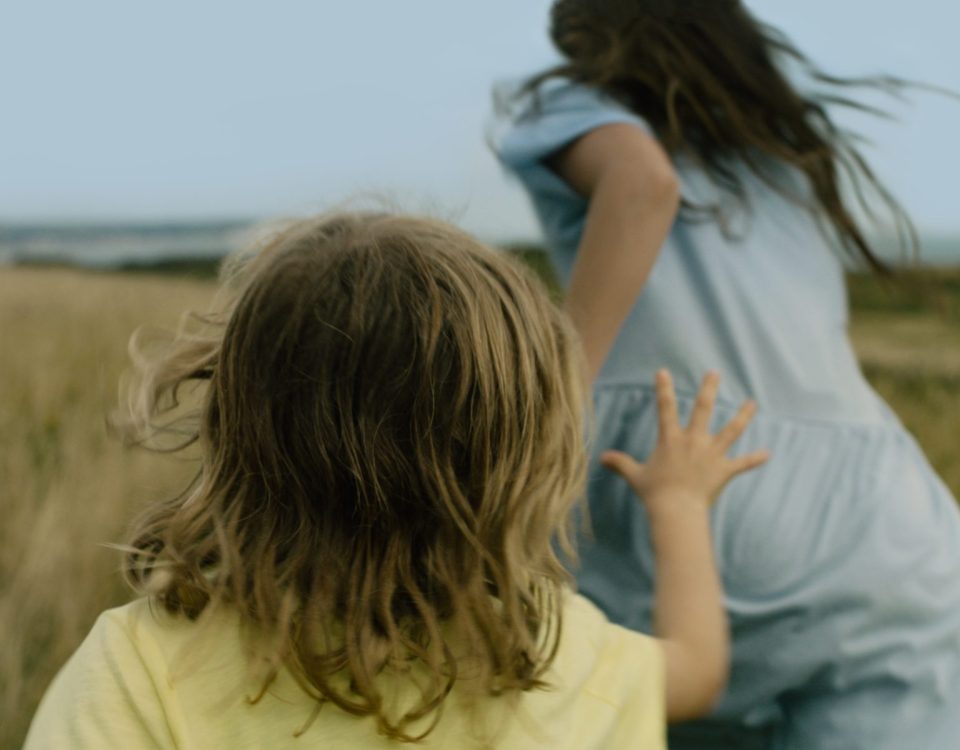 Two girls play chase in the grass, one younger wearing yellow, one older wearing blue, with a pale blue sky behind them.